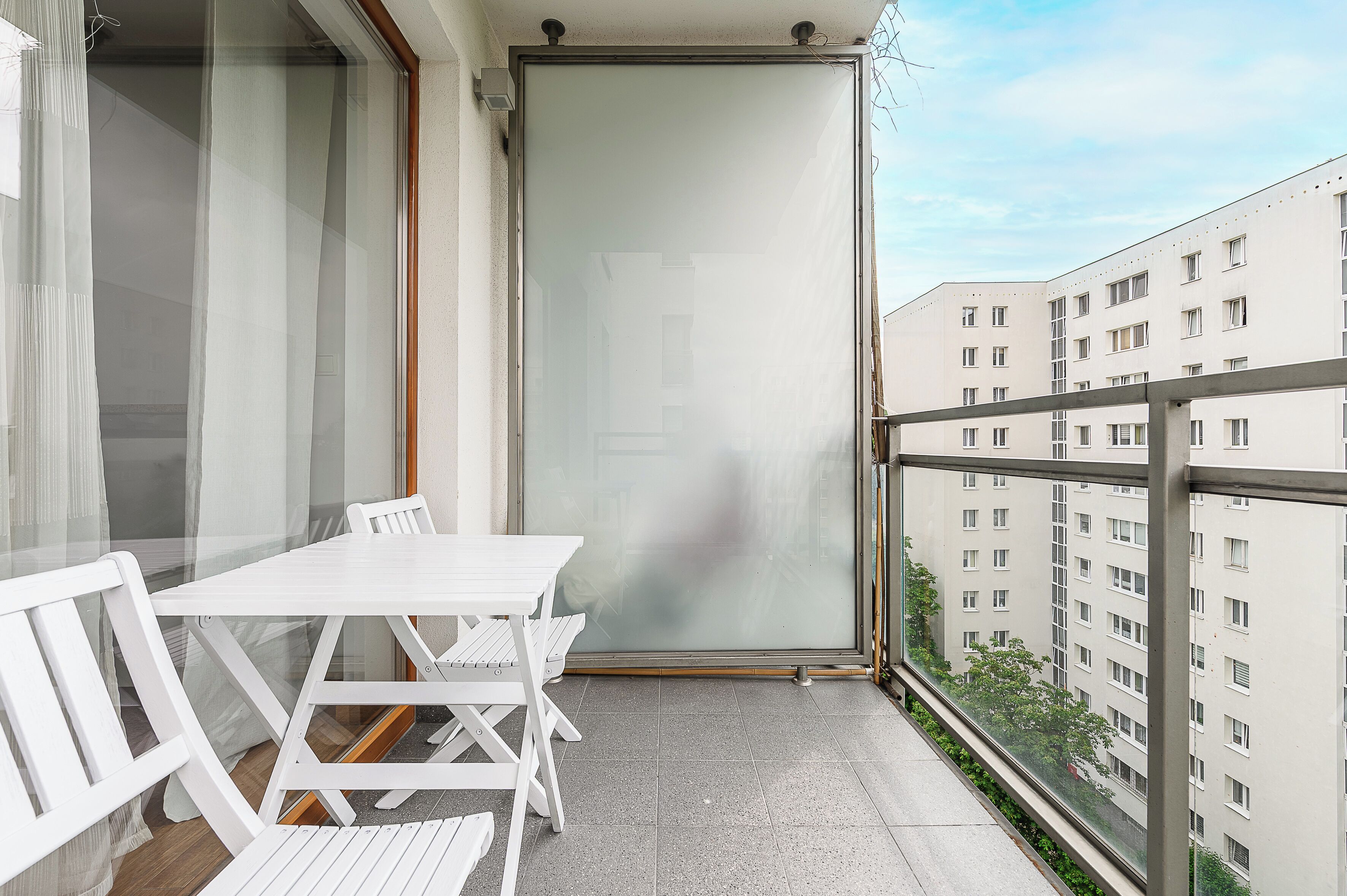 Ein Balkon mit weißem Tisch und Stühlen, einer Schiebetür aus Glas und Blick auf angrenzende Gebäude.