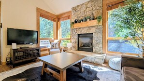 A cozy living room with a stone fireplace, a flat-screen TV, a wooden table, and a large window offering a view of trees.