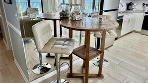 Een moderne keuken met een ronde houten tafel en twee barkrukken, met uitzicht op de oceaan door grote ramen, en een balkon met een stoel.