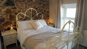 Un lit adossé à un mur en pierre, une table de chevet avec une lampe et une fenêtre avec des rideaux.