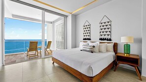 A bedroom with a large bed, a nightstand, a lamp, two wall hangings, and a view of the ocean through a sliding glass door. - Beachfront 2BR - Pools & Scenic Balcony Views (Puerto Vallarta)
