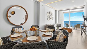A modern dining area with a round glass table, woven chairs, and a view of the ocean through large windows. - Beachfront 2BR - Pools & Scenic Balcony Views (Puerto Vallarta)