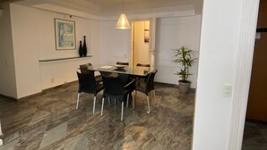 Un comedor con suelo de mármol, una mesa de vidrio, sillas negras, una lámpara colgante, un cuadro en la pared y una planta en maceta.