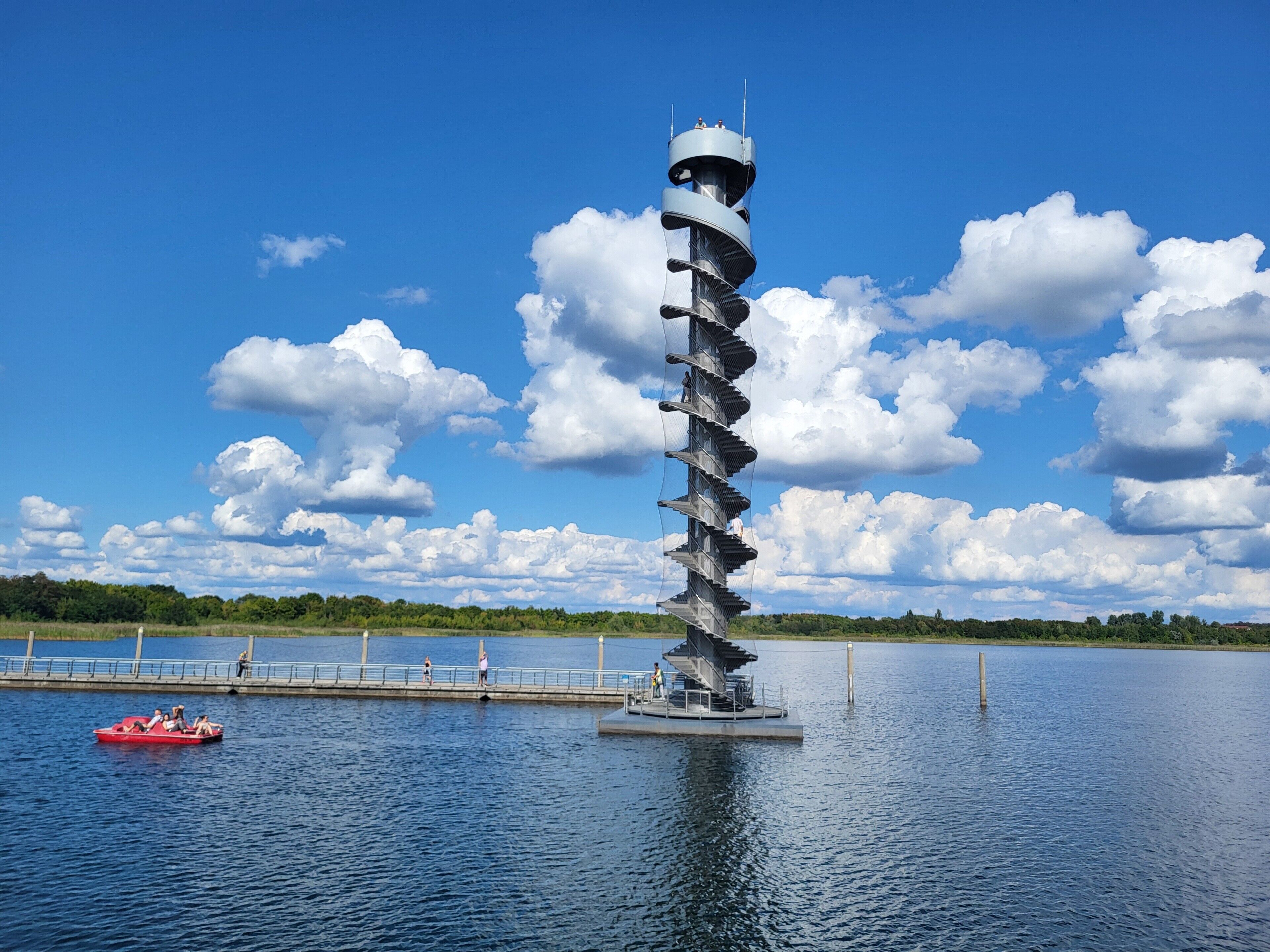 Eine spiralförmige Turmstruktur auf dem Wasser, mit blauem Himmel und weiter, grüner Landschaft im Hintergrund.