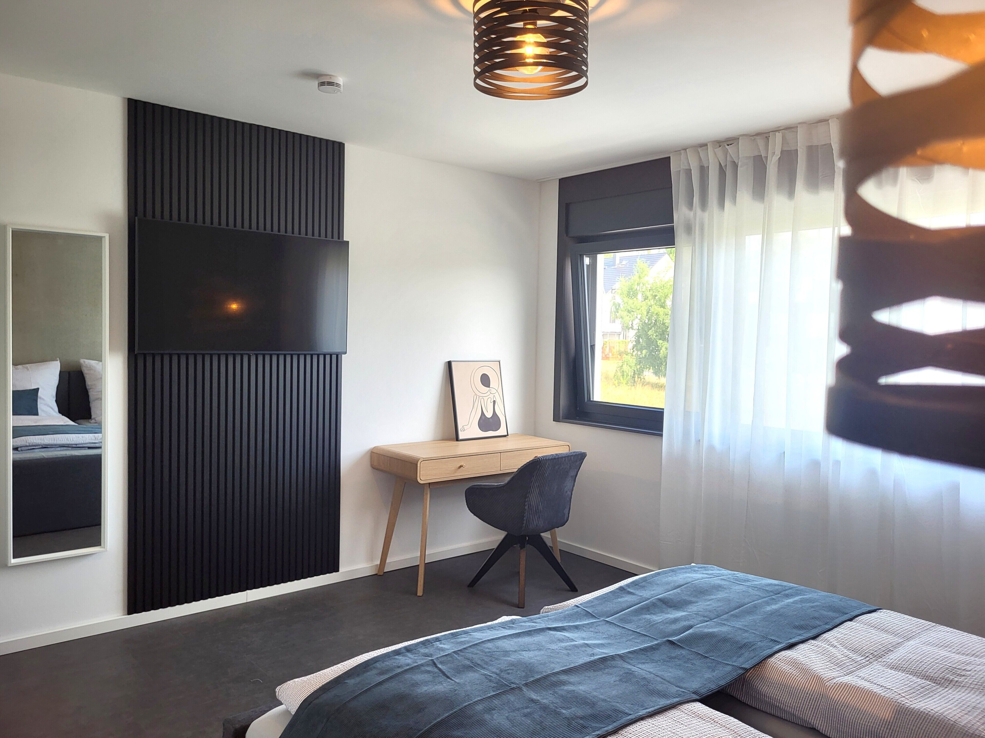 A bedroom with a bed, a desk, a chair, a window with curtains, and a wall-mounted TV.