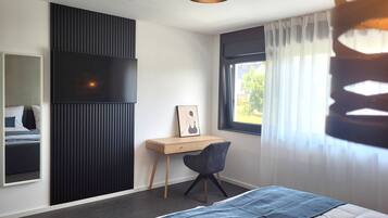 A bedroom with a bed, a desk, a chair, a window with curtains, and a wall-mounted TV.