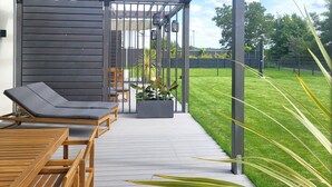 Une terrasse couverte, meublée de mobilier en bois, agrémentée d’un jardinier avec des plantes, et offrant une vue sur une pelouse bien entretenue.