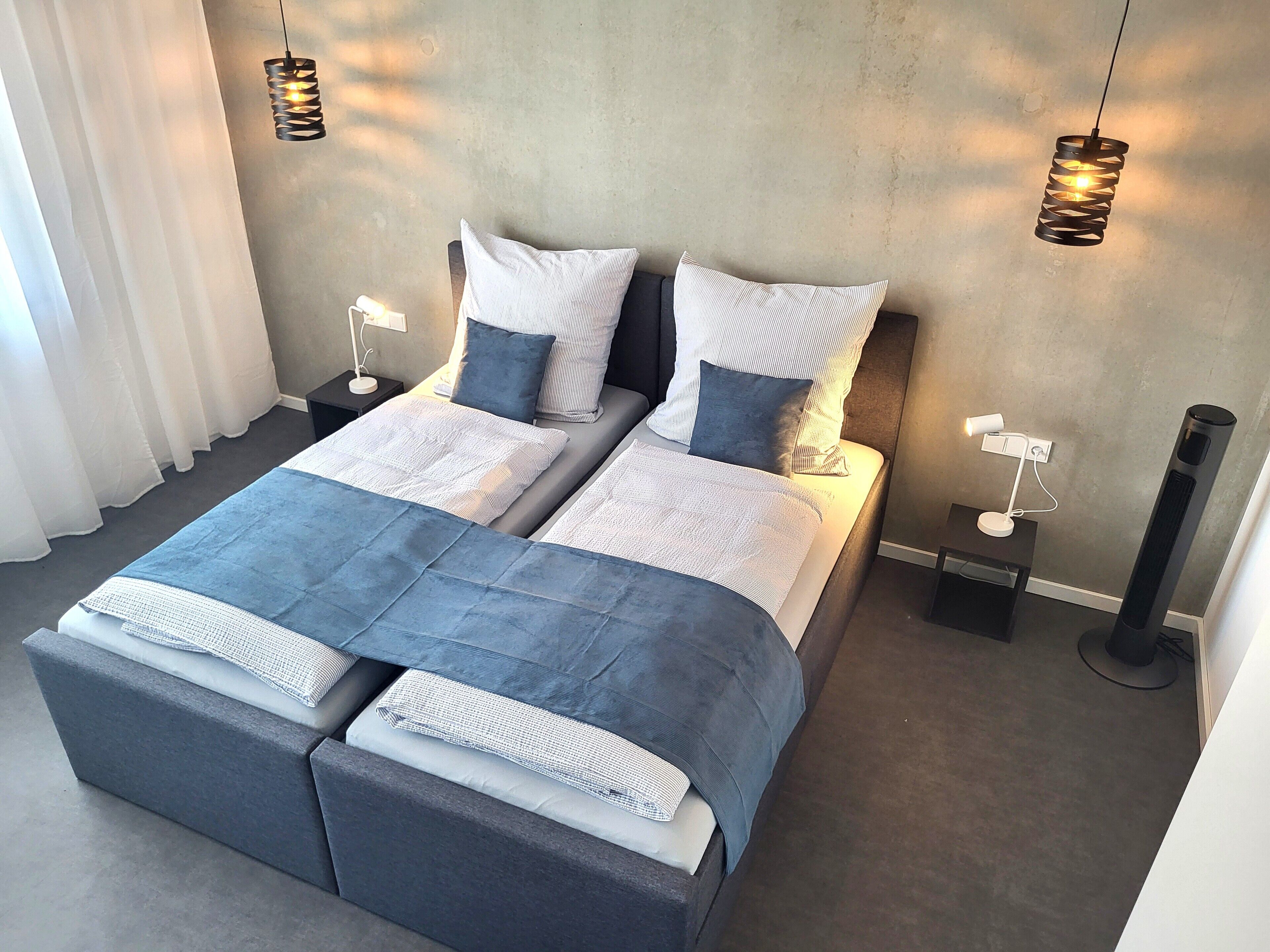 Ein Doppelbett mit grauem Kopfteil, zwei Nachttische mit Lampen sowie ein großes Fenster mit weißen Vorhängen.