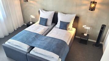 Ein Doppelbett mit grauem Kopfteil, zwei Nachttische mit Lampen sowie ein großes Fenster mit weißen Vorhängen.