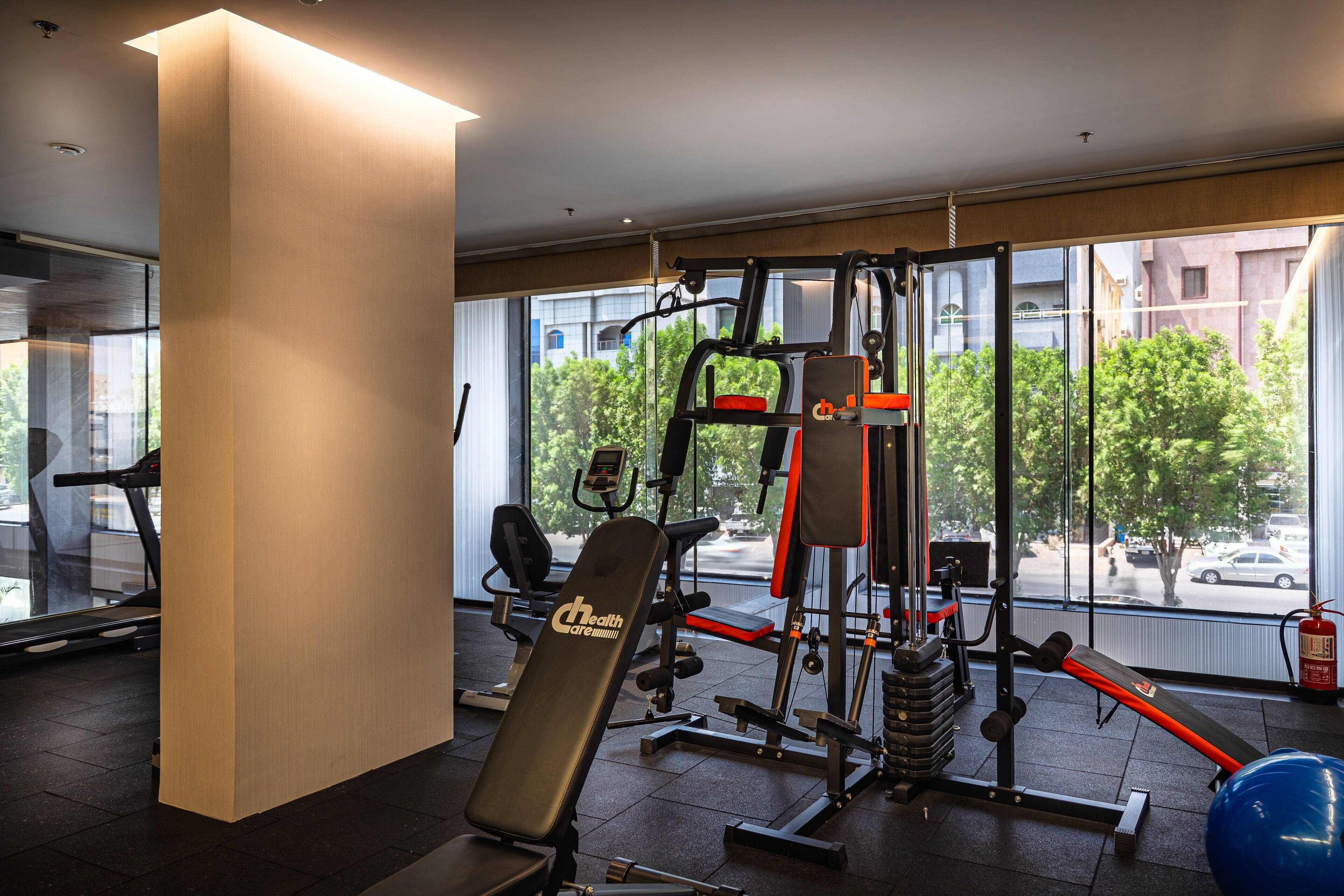 Un gimnasio con equipo de ejercicio, una banca y un ventanal con vistas a árboles y edificios.