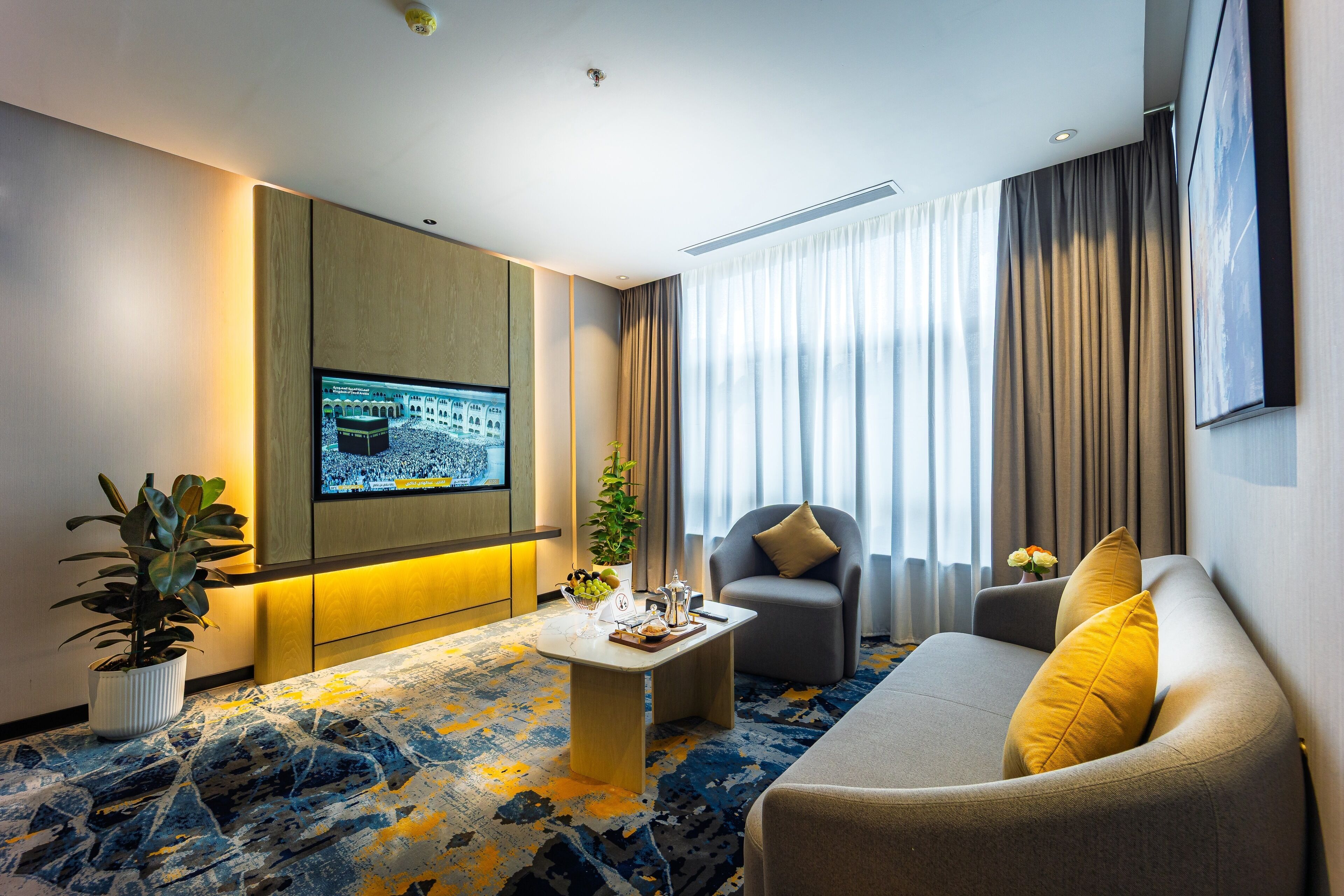 A modern hotel room with a flat-screen TV mounted on the wall, a comfortable sofa with yellow cushions, a small coffee table, and a patterned carpet.