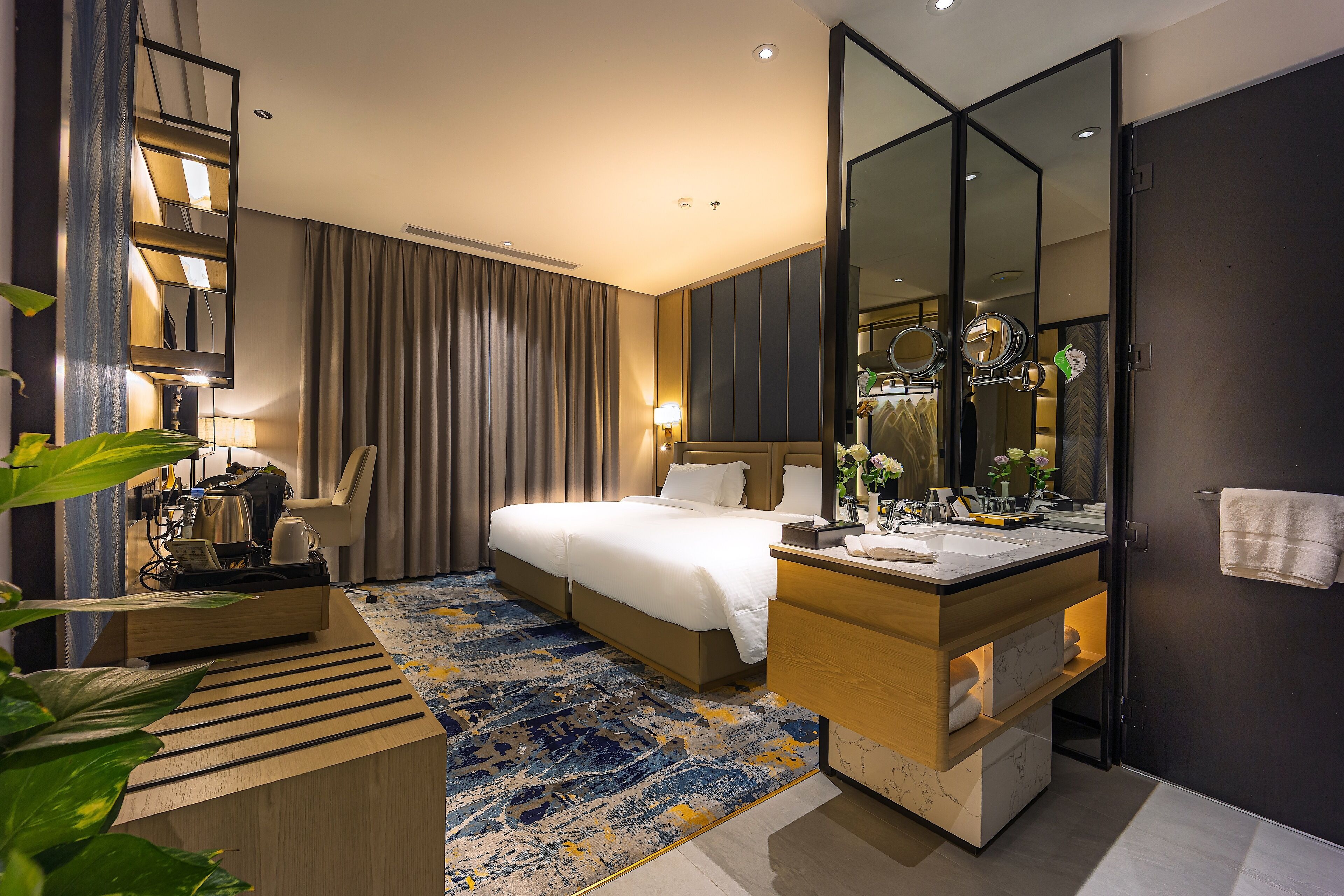 A modern hotel room with a large bed, a wooden desk, a bathroom with a mirror and sink, and a large mirror on the wall.