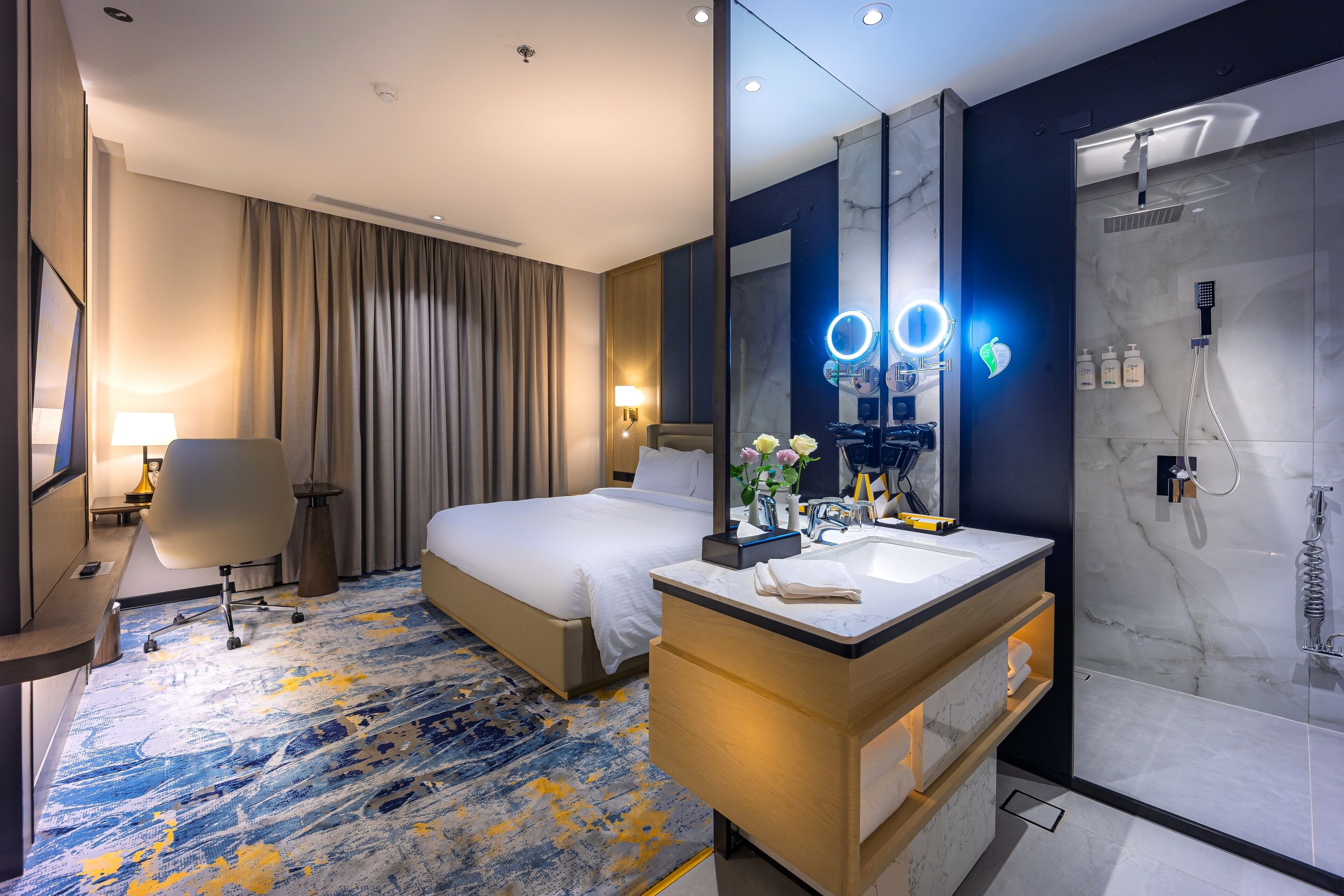 A hotel room with a bed, a desk, a chair, a bathroom with a shower, and a large mirror.