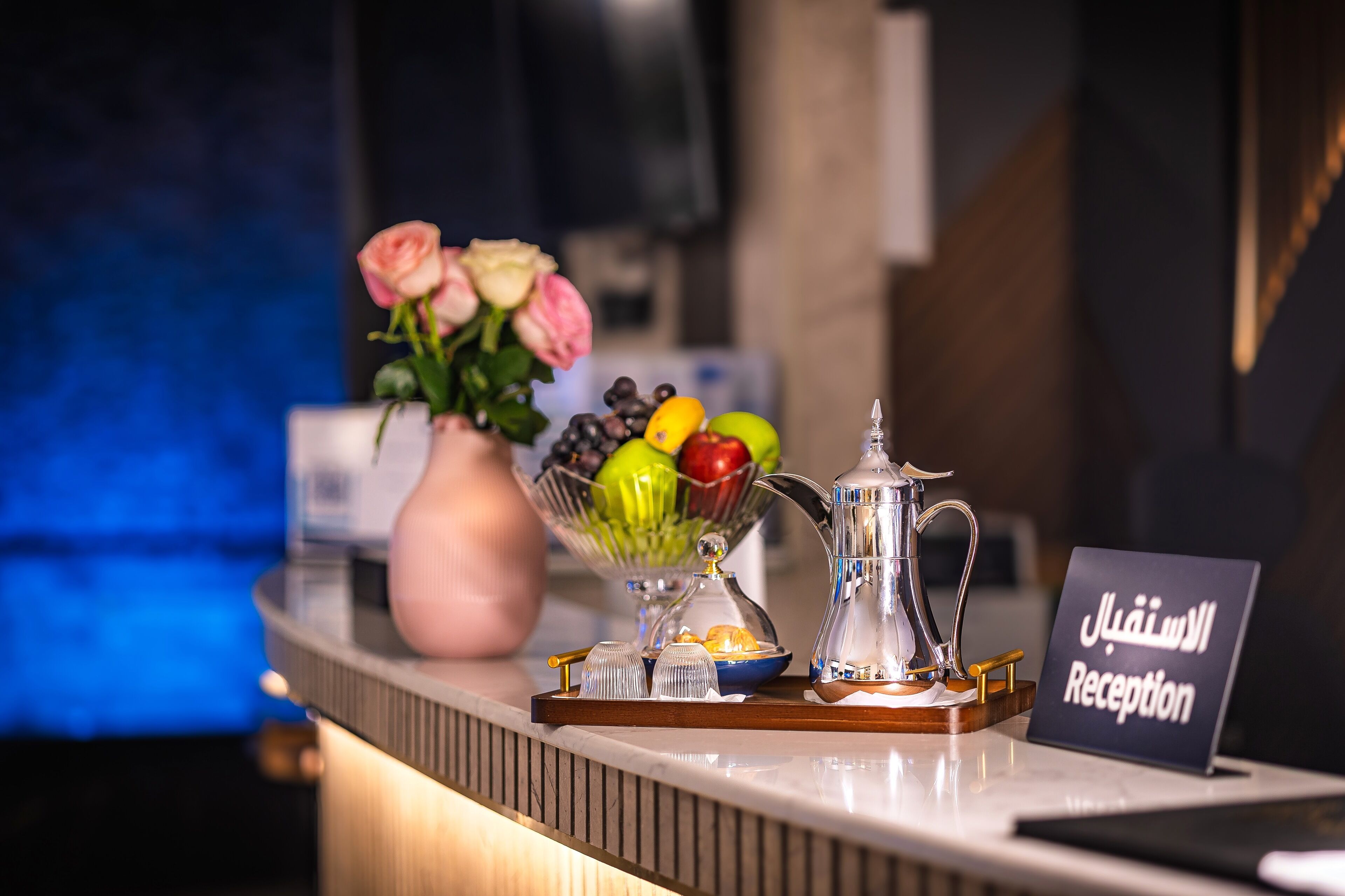 Ein Empfangsschalter mit einem Schild, das 'Rezeption' auf Arabisch zeigt, eine Vase mit rosa Rosen, eine Obstschale und eine Kaffeekanne.