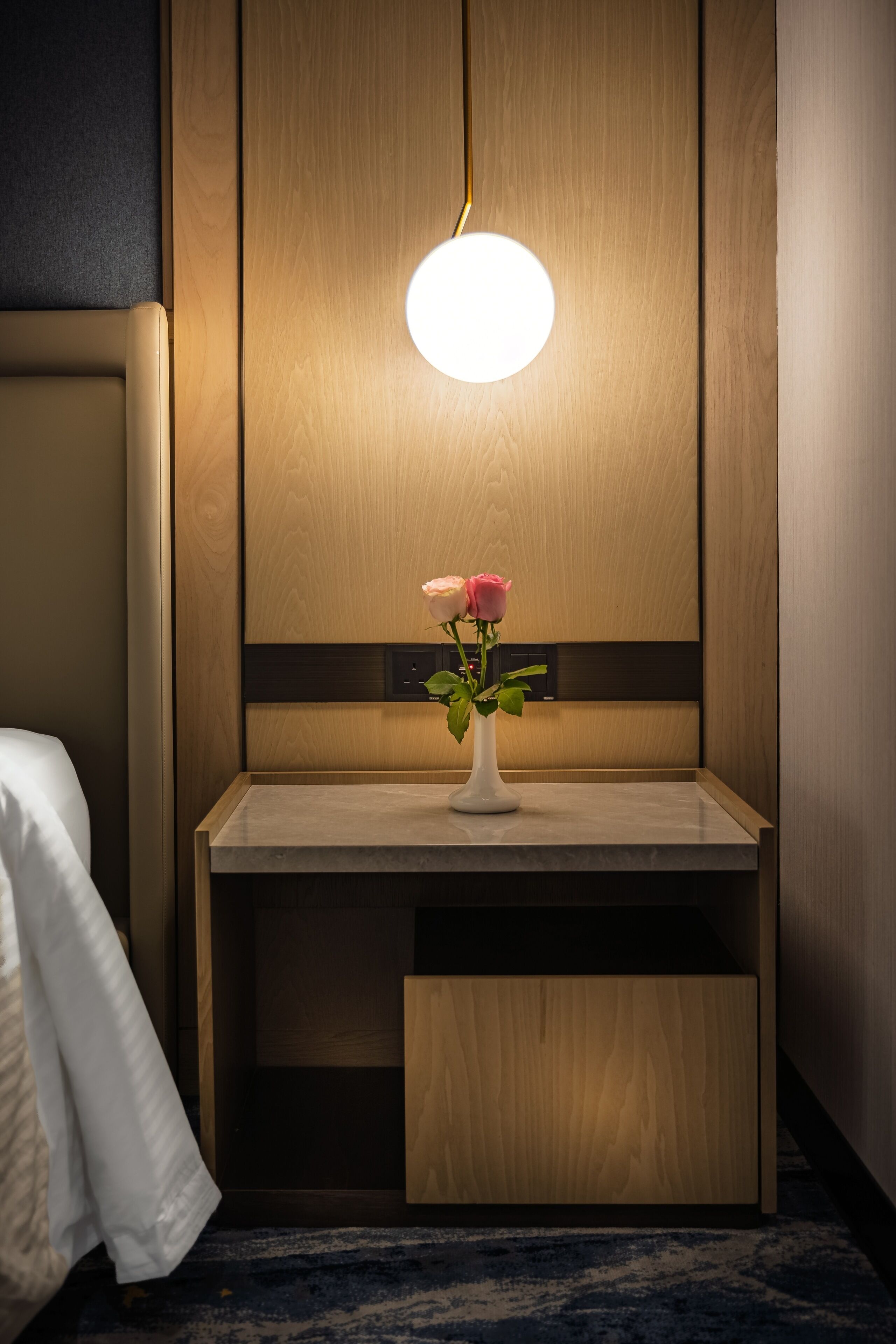 Habitación de hotel con pared de paneles de madera, una mesita de noche con un jarrón de flores y una lámpara de techo redonda.