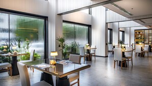 Un comedor moderno con mesas y sillas de vidrio, ventanas amplias y plantas de interior.