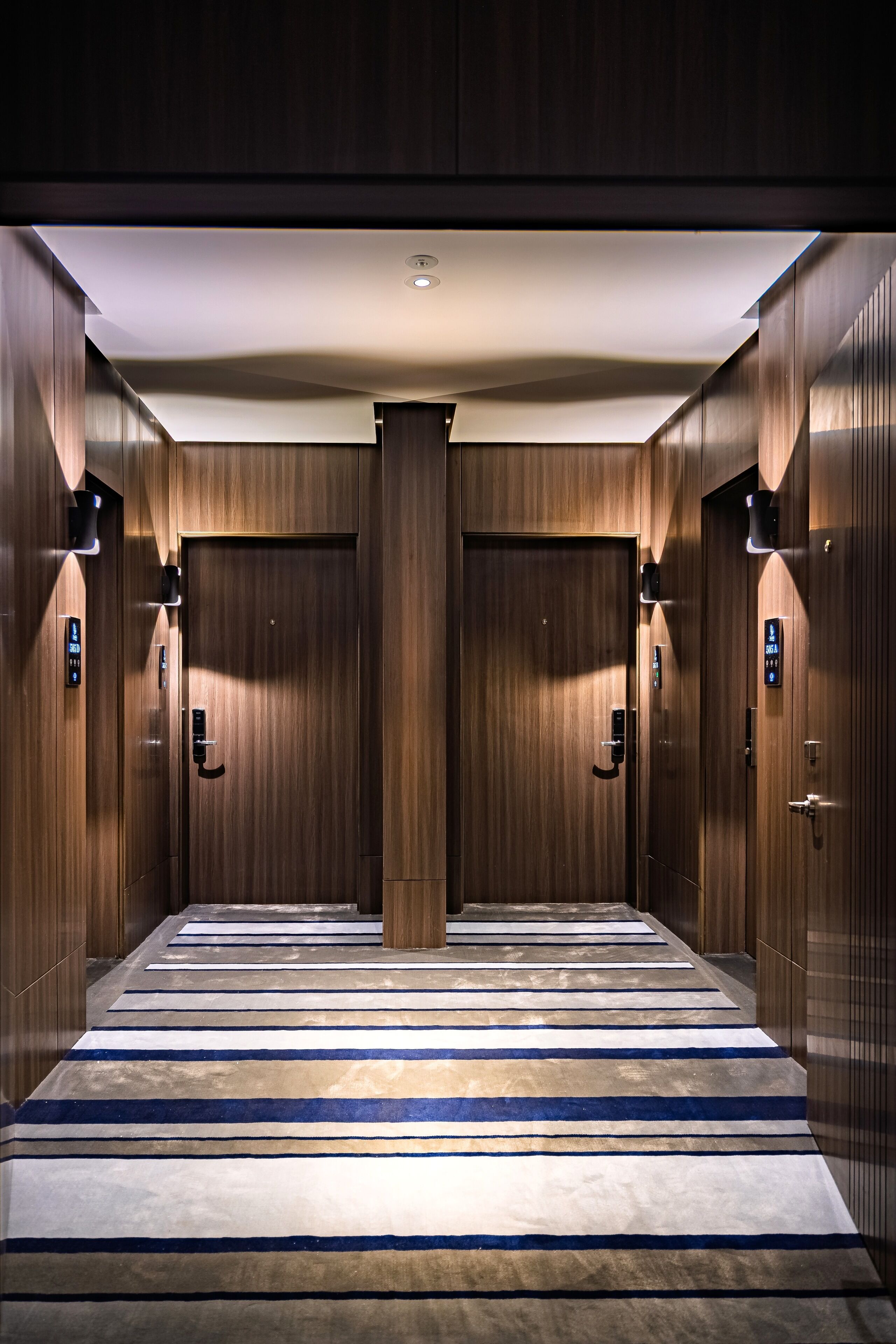 Un couloir aux murs en bois et au tapis rayé, qui mène à une porte dotée d’un écran numérique.