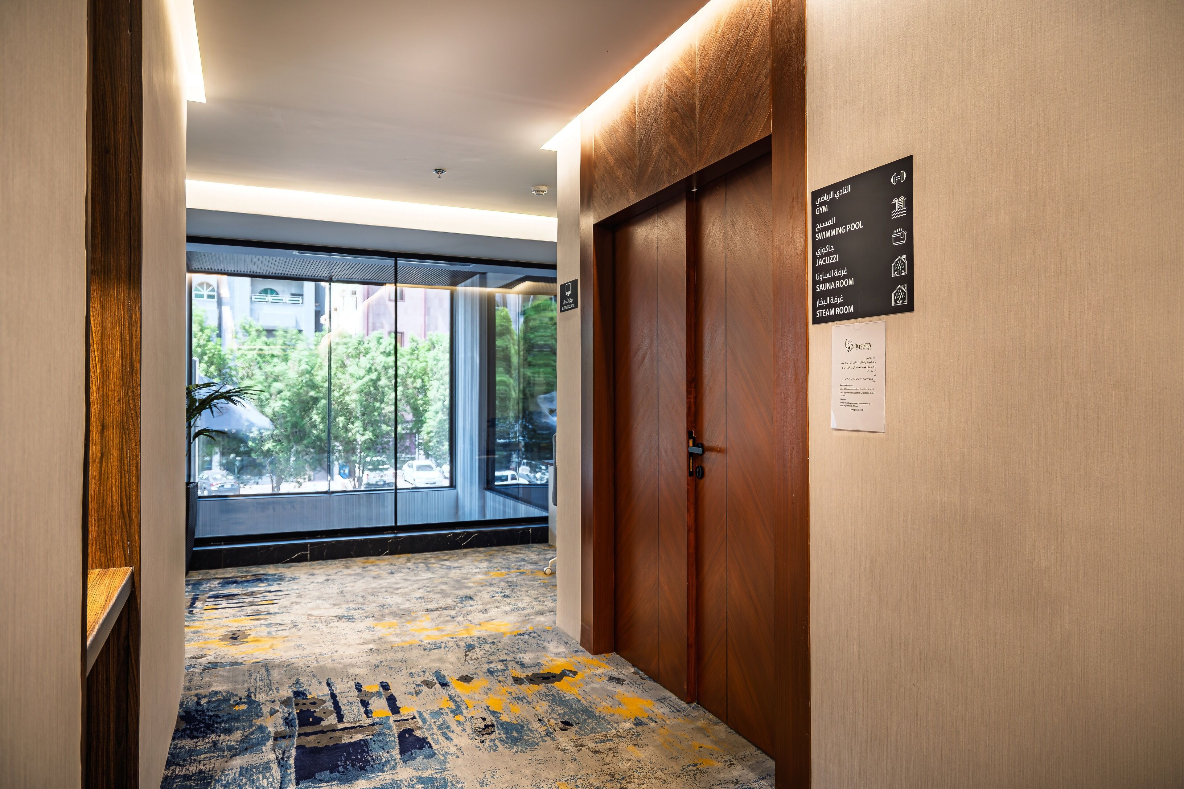 A hotel corridor with a wooden door, a sign listing amenities, and a view through a glass door.