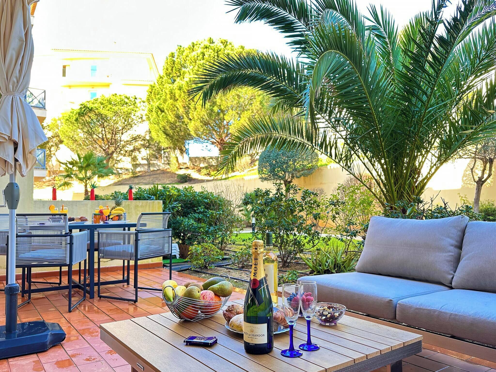 Une terrasse extérieure avec une table en bois, une bouteille de champagne et un saladier de fruits.
