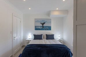 A bedroom with a bed, two bedside tables, a wall-mounted painting of a whale tail, and a door. - Luxury Oceanfront Retreat – Whaleview Villa, Klein Slangkop, Kommetjie (Cape Town)