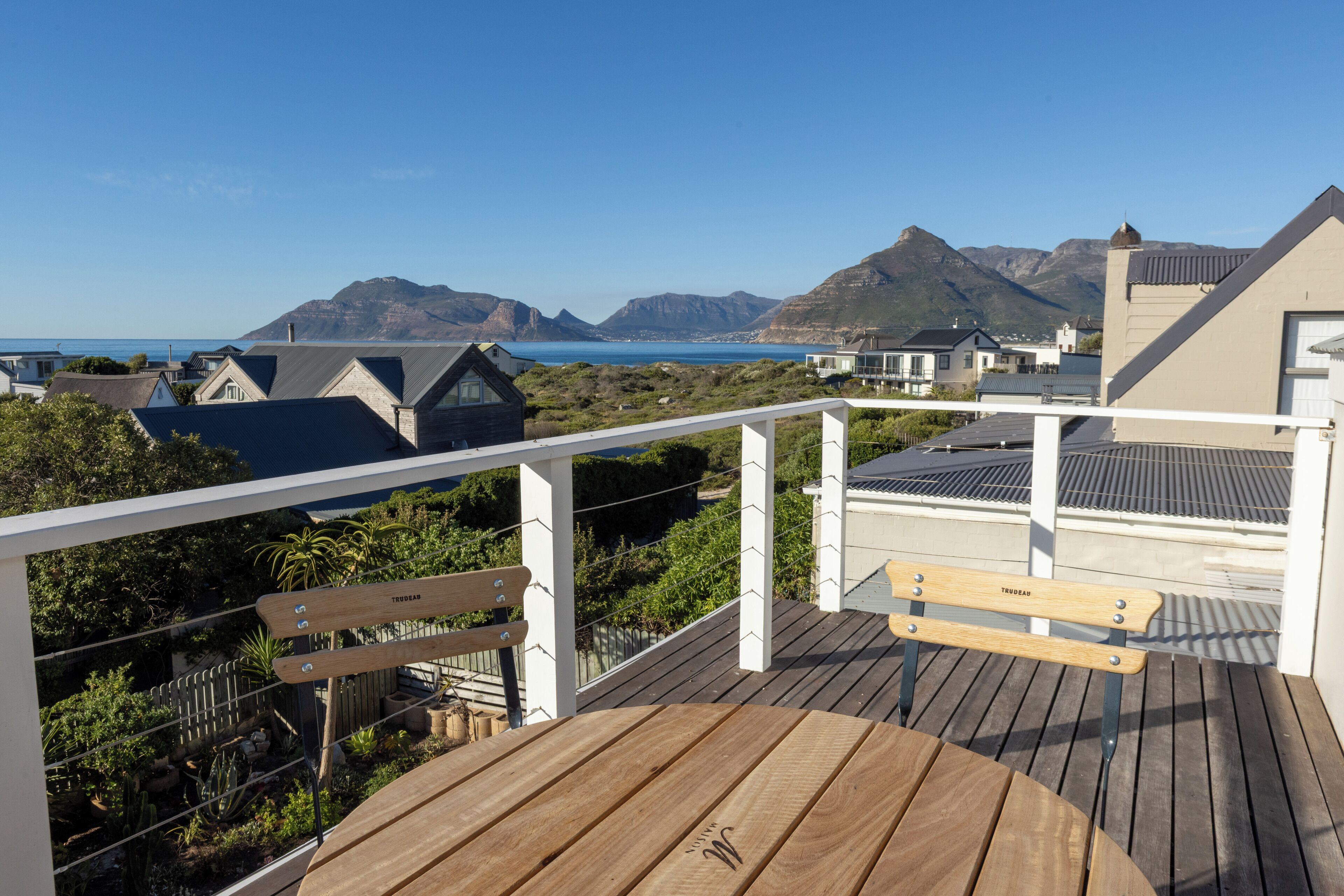 Een houten terras met een tafel en banken, met uitzicht op een kustlandschap met bergen en huizen.