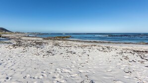 Ein Strand mit weißem Sand und Seetang, ein klarer blauer Himmel und in der Ferne felsige Formationen.