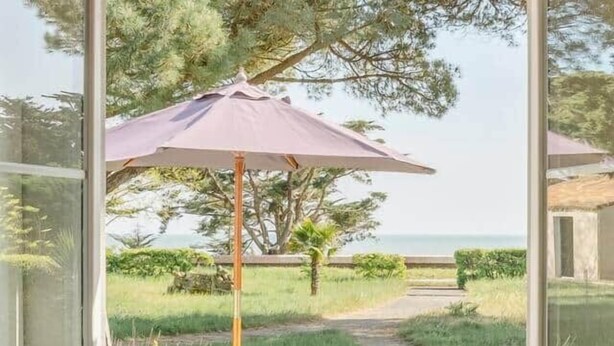 A dining table set with plates and glasses, a vase with flowers, and a view of a grassy area with trees and a path. - Villa Kenavo - Family house with sea view (Île-d'Aix)