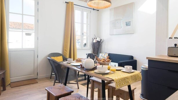 A dining area with a table set for breakfast, a sofa, and a view of the street outside. - La Miniardise - House in the village (Île-d'Aix)