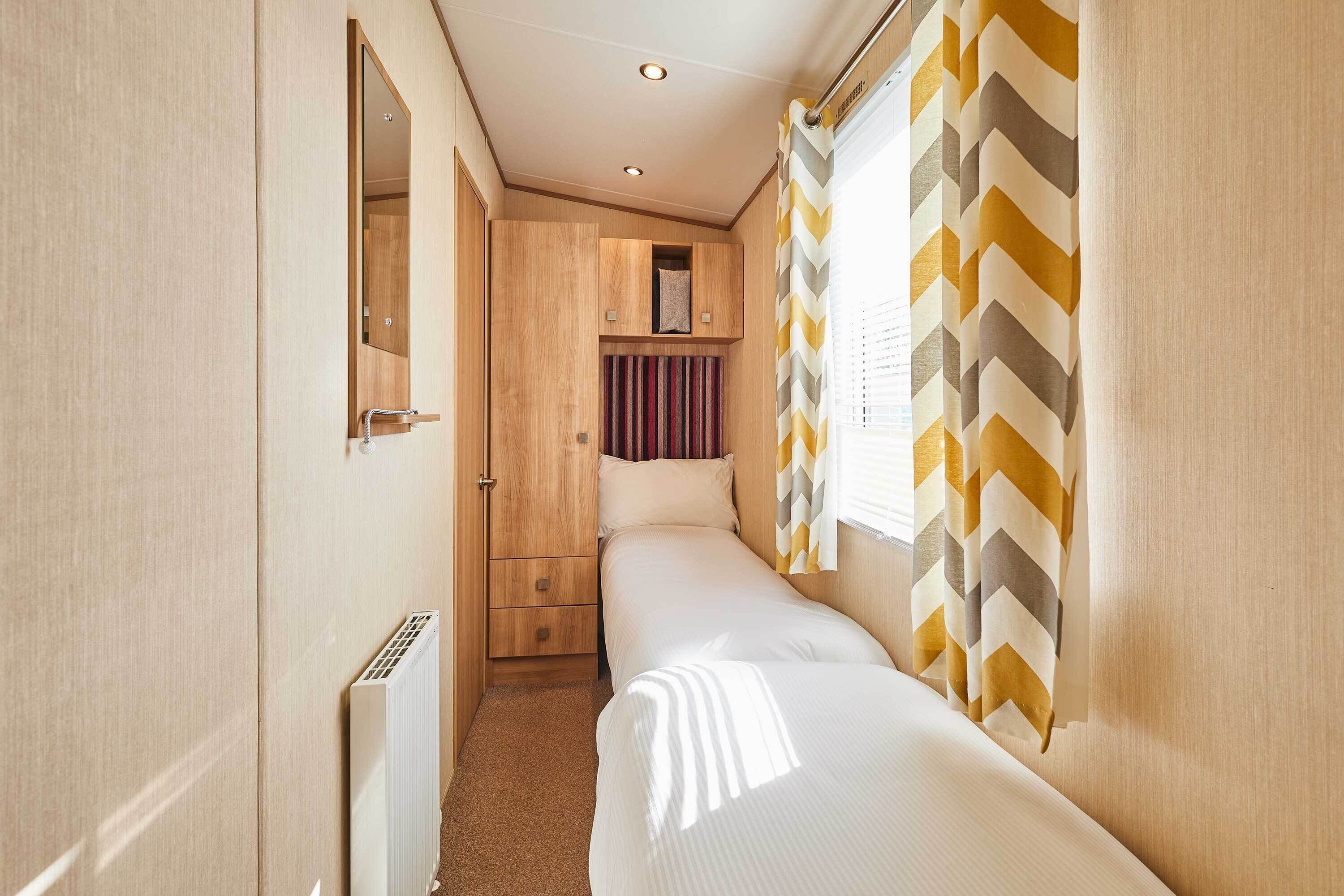 Um quarto pequeno e bem iluminado com uma cama de solteiro, um armário de madeira, um radiador e cortinas com padrão em chevron.