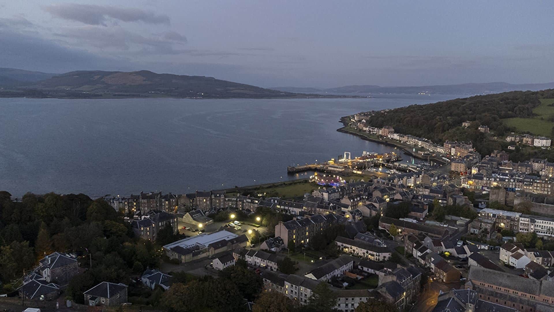 A coastal town with a mix of residential and commercial buildings, a marina, and a view of the sea and hills in the distance.