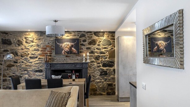 A cozy living room with a stone wall, a fireplace, and a framed picture of a bull. There is a dining area with a table and chairs, and a modern light fixture above. - Scalpsie Luxury Apartment (Rothesay)