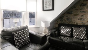 A modern living room with a black leather sofa, patterned cushions, a side table with a lamp, and a framed artwork on the wall.