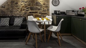 A cozy dining area with a stone wall, a black leather sofa with patterned cushions, a white dining table with chairs, and a kitchen area with a microwave and coffee maker.