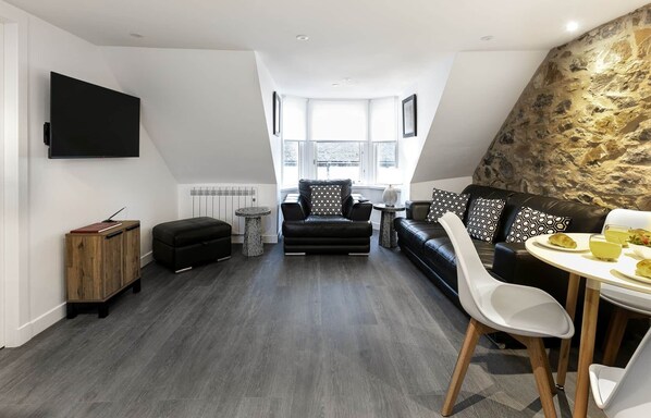 Un salon moderne de style industriel, avec un canapé en cuir noir, une table à manger blanche et un mur d’accent en pierre.