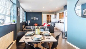 A modern living room with a dining area, featuring a sofa, a table set for two, and a kitchen in the background.