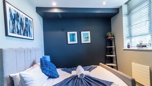 A modern hotel room with a bed, a large window with blinds, a wall-mounted shelf with decorative items, and a framed artwork above the bed.