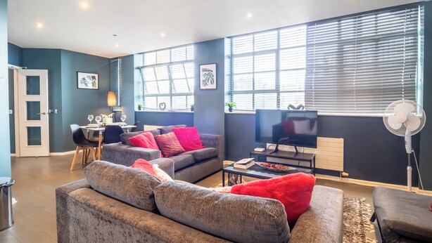 A modern living room with grey sofas, red cushions, a dining area, a TV, and a large window with blinds.