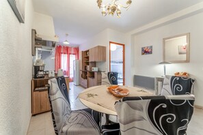 A dining area with a round table, chairs, a chandelier, and a kitchen in the background.
