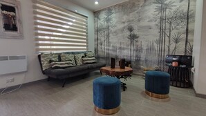 A living room with a grey sofa, a round coffee table, two blue ottomans, a wall-mounted TV, and a forest-themed mural. - Oceana Suites Los Nostros (Pucon)