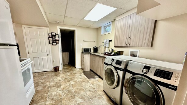 A laundry room with a washing machine, dryer, and a microwave. There is a door leading to another room and a window above the sink. - Beautiful Suite with Fenced yard + in suite Kitchen and Laundry (St. Albert)