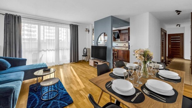 Ein modernes Wohnzimmer mit einem blauen Sofa, einem Esstisch für vier Personen und einer Küche im Hintergrund.