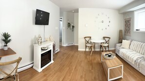 Uma sala de estar com sofá branco, mesa de centro de madeira, televisão fixada na parede, lareira e um espaço de jantar com duas cadeiras e uma pequena mesa.