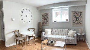 Uma sala de estar com um sofá branco, duas cadeiras de madeira, uma mesa redonda pequena e um relógio de parede.