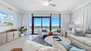 En rummelig stue med store skydedøre til en balkon med udsigt over stranden. Der er ventilator i loftet, et fladskærms-tv monteret på væggen og et sofabord med en bakke.