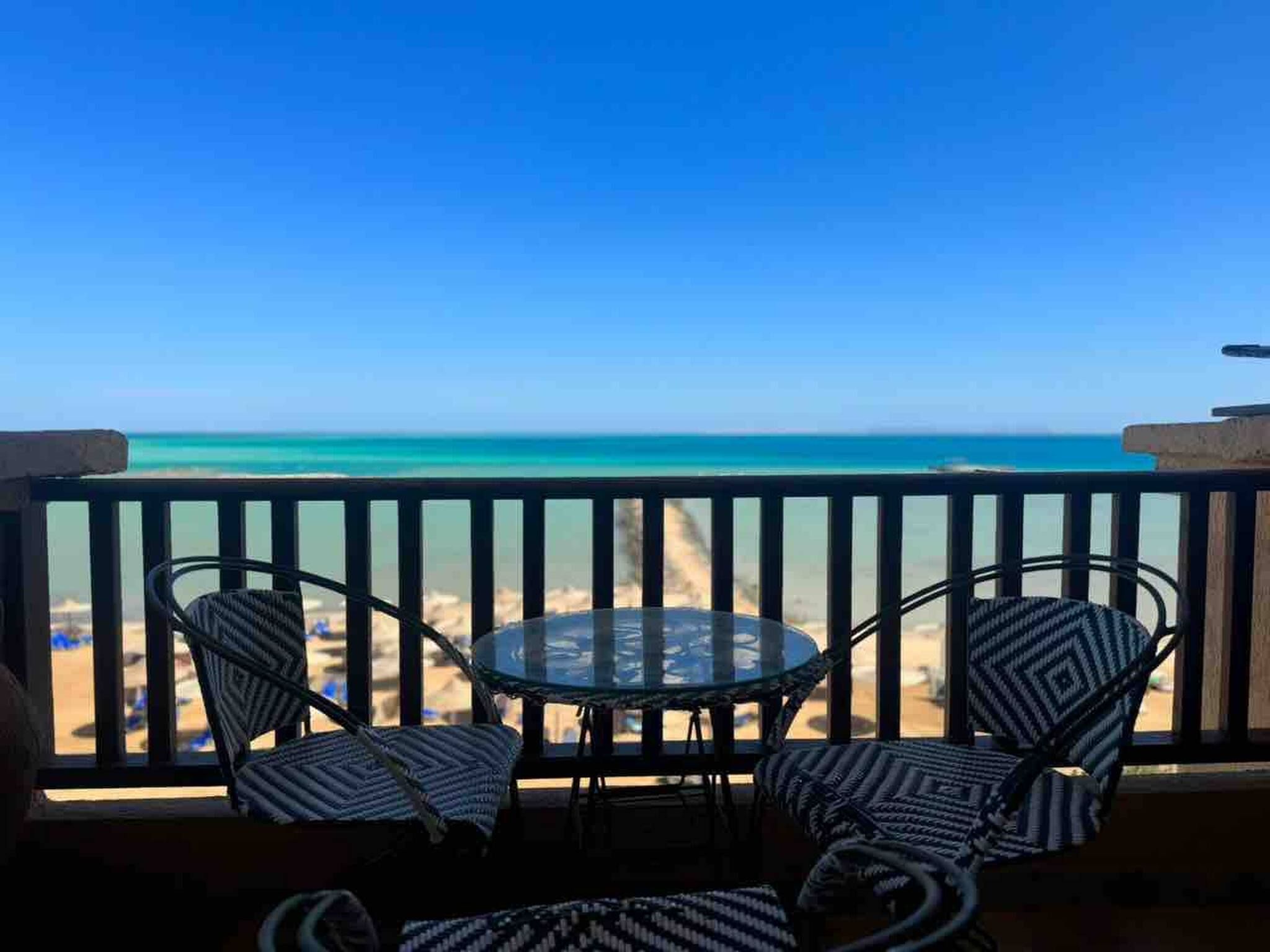 A balcony with a glass table and two chairs, overlooking a beach with clear blue water.