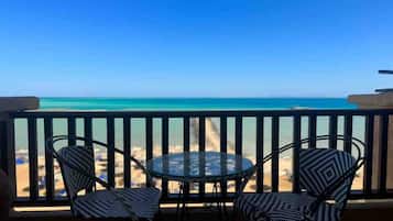 A balcony with a glass table and two chairs, overlooking a beach with clear blue water.