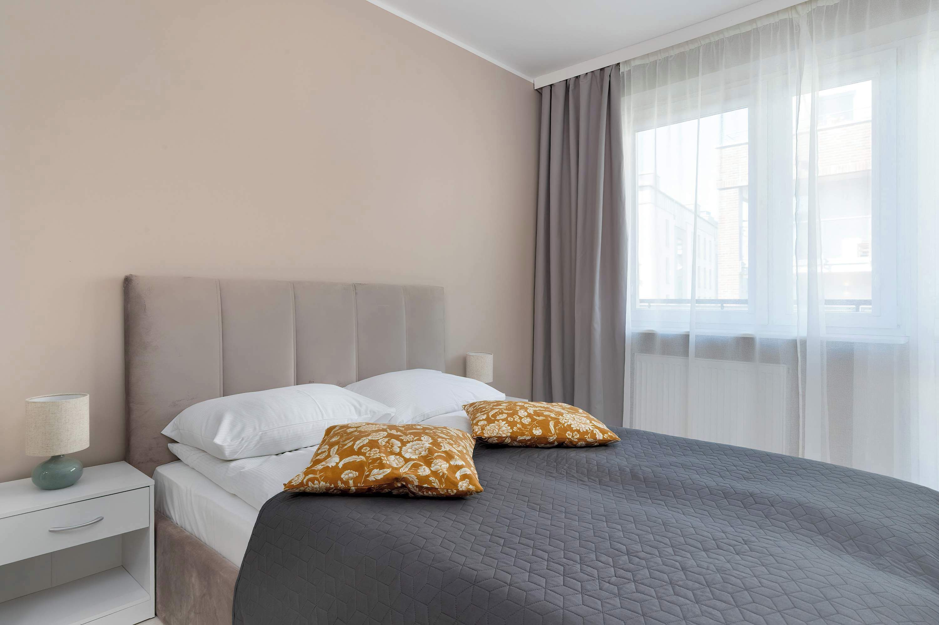 Un lit bien fait, recouvert d’une couette grise, avec deux oreillers blancs et deux oreillers à motifs. On aperçoit une table de chevet avec une lampe et un tiroir blanc.