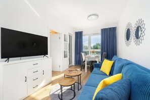 A modern living room with a flat-screen TV, a blue sofa, a wooden coffee table, and a dining area with a table and chairs.