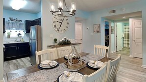 A dining area with a wooden table set for four, surrounded by white chairs. A large wall clock and a chandelier are visible above the table. - Prime Pines | Great Views, Great Location, Resort Amenities (Pigeon Forge)
