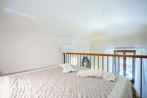 Un dormitorio con cama, almohadas, una ventana con cortinas y barandilla de balcón.